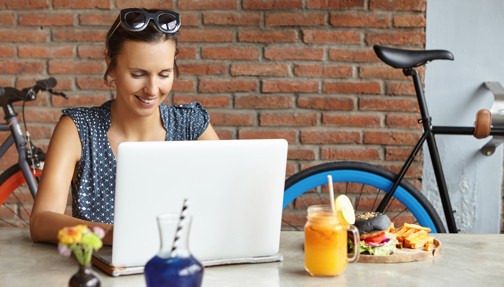 Attractive woman photographer retouching pictures using photo editor, having lunch, sitting in front of generic laptop. Student girl studying online on notebook, smiling joyfully while making progress filipino photo retoucher